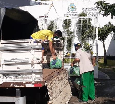 dois homens descarregando grandes sacolas de caminhão na porta da secretaria regional um